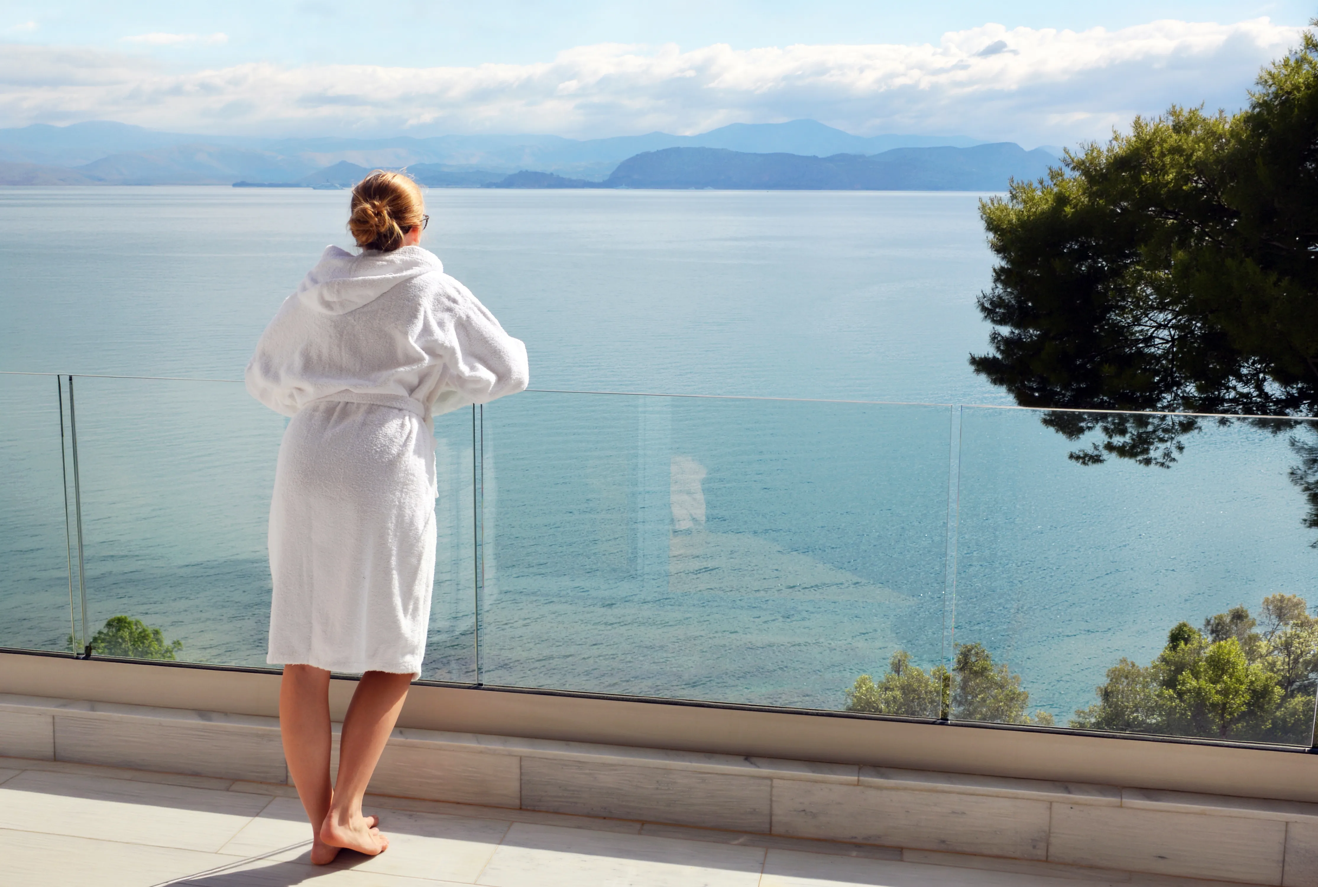 Gast im Bademantel an der Glasbrüstung des Balkons mit Blick über den ruhigen Golf und die Berge