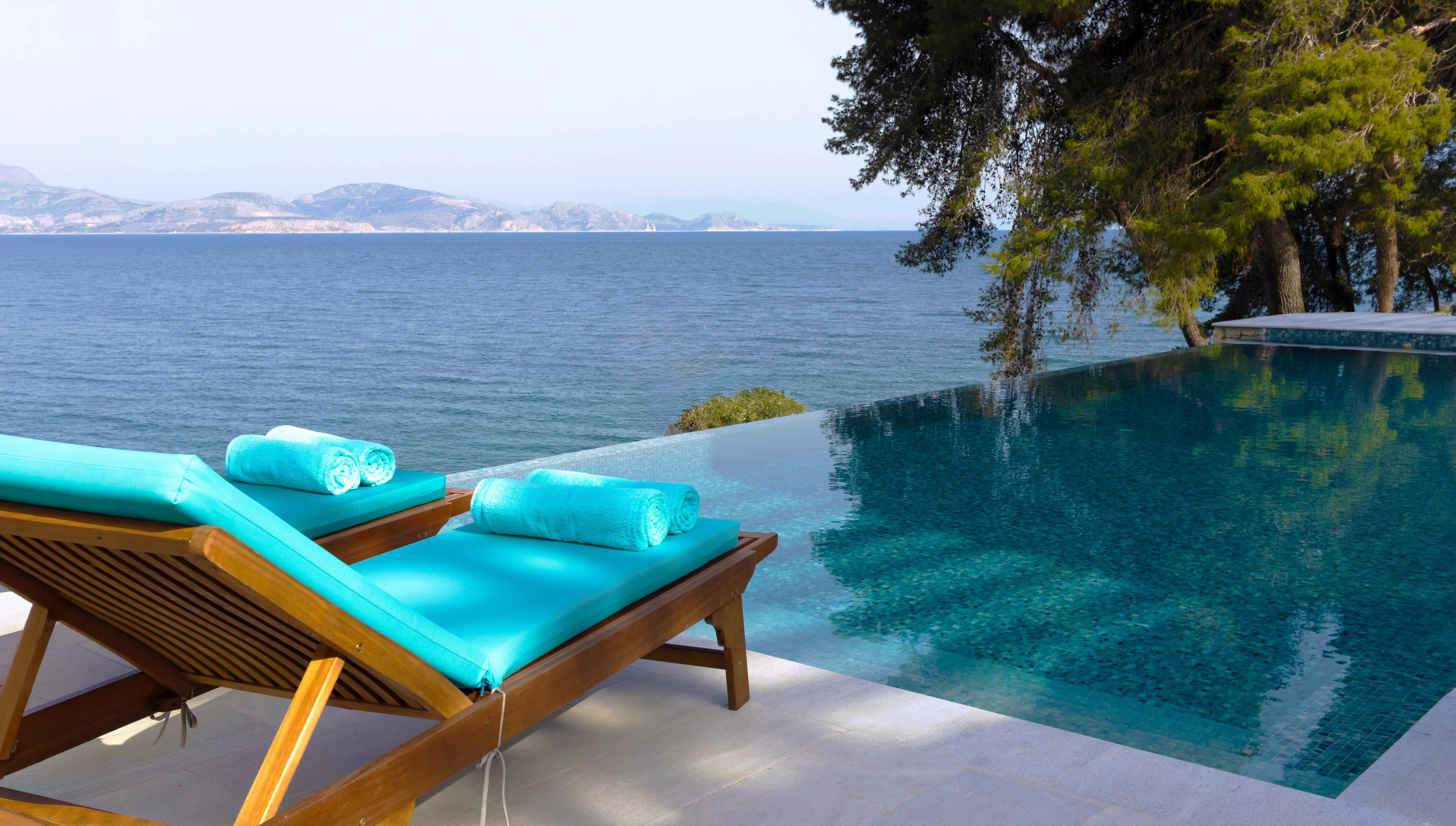 Holzliegen mit türkisfarbenen Auflagen am Infinity-Pool über dem Meer
